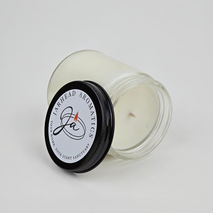 A white candle, The Lumberjack Soy Wax Candle by Jarhead Aromatics, rests in a clear glass jar with its black logo lid nearby, suggesting a masculine cedar, oak & mahogany scent inspired by a woodland cabin sanctuary.