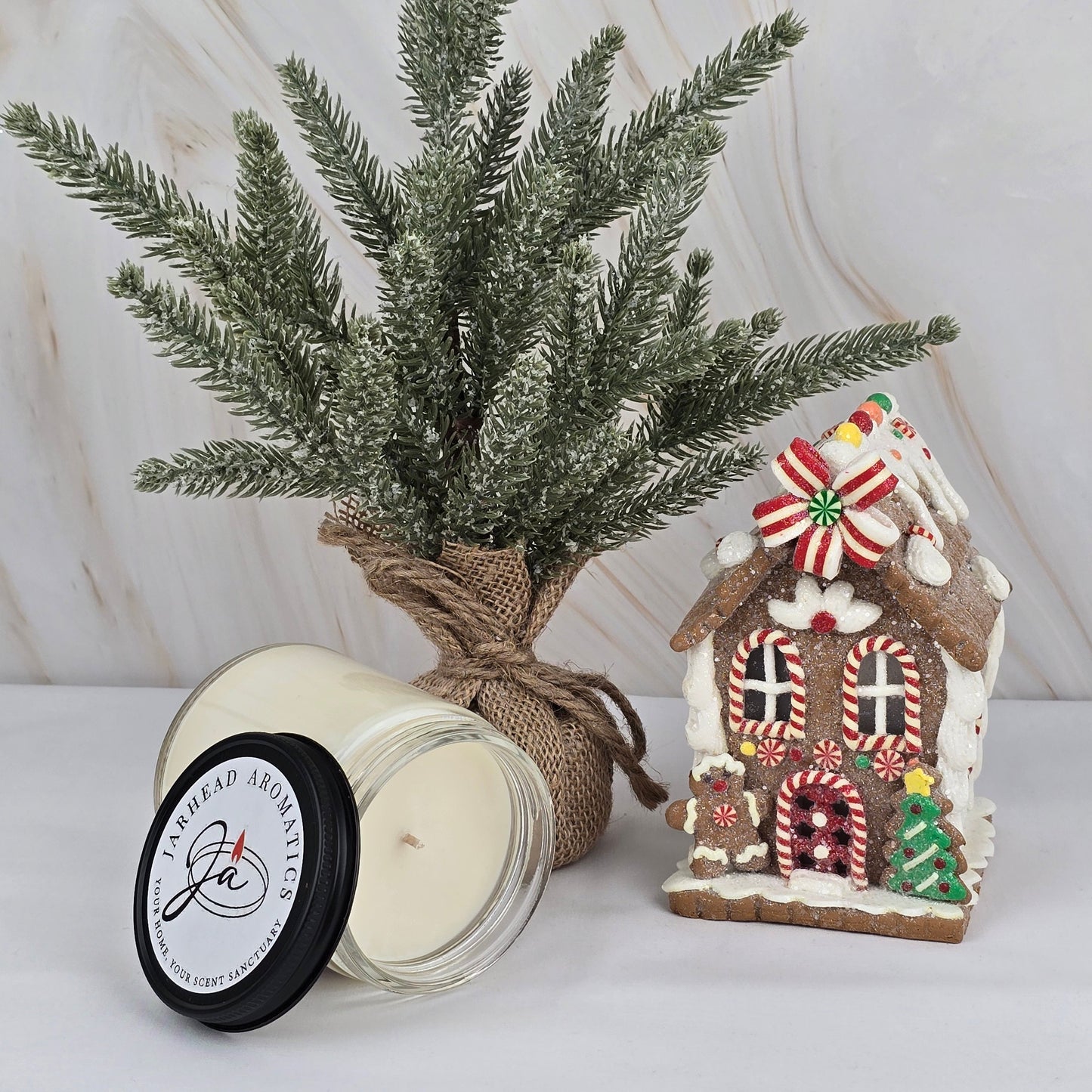 A frosted faux pine tree in burlap sits next to a decorated gingerbread house and Jarhead Aromatics soy wax candle from the Winter Collection, all arranged on a white surface with a marble backdrop.