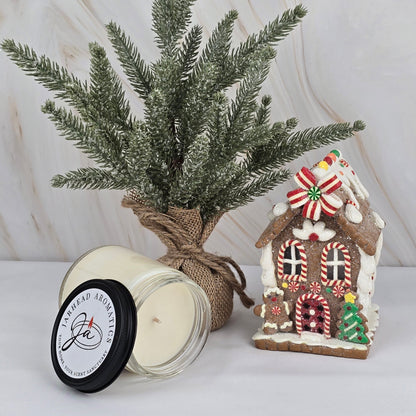A frosted faux pine tree in burlap sits next to a decorated gingerbread house and Jarhead Aromatics soy wax candle from the Winter Collection, all arranged on a white surface with a marble backdrop.