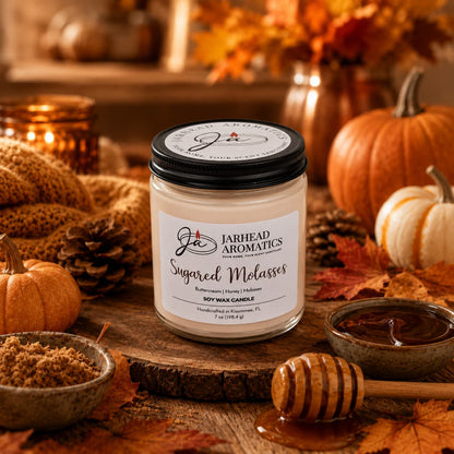 A Sugared Molasses Soy Wax Candle by Jarhead Aromatics sits on a rustic table surrounded by pumpkins, autumn leaves, a bowl of brown sugar, a honey dipper with honey, and a softly glowing candle—evoking the warmth and coziness of fall. This handmade Brown Sugar, Honey, Cinnamon & Molasses Fall Candle fills the room with a rich gourmand fragrance that perfectly captures the essence of autumn.
