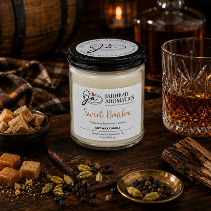 A Jarhead Aromatics Sweet Bourbon Soy Wax Candle | Warm Bourbon, Brown Sugar & Oak Candle rests on a wooden table alongside whiskey, a glass of bourbon, sugar cubes, spices, and a checked cloth, creating a warm, rustic ambiance. This hand-poured soy candle offers a long-lasting blend of bourbon, brown sugar, and oak that perfectly complements the inviting setting.