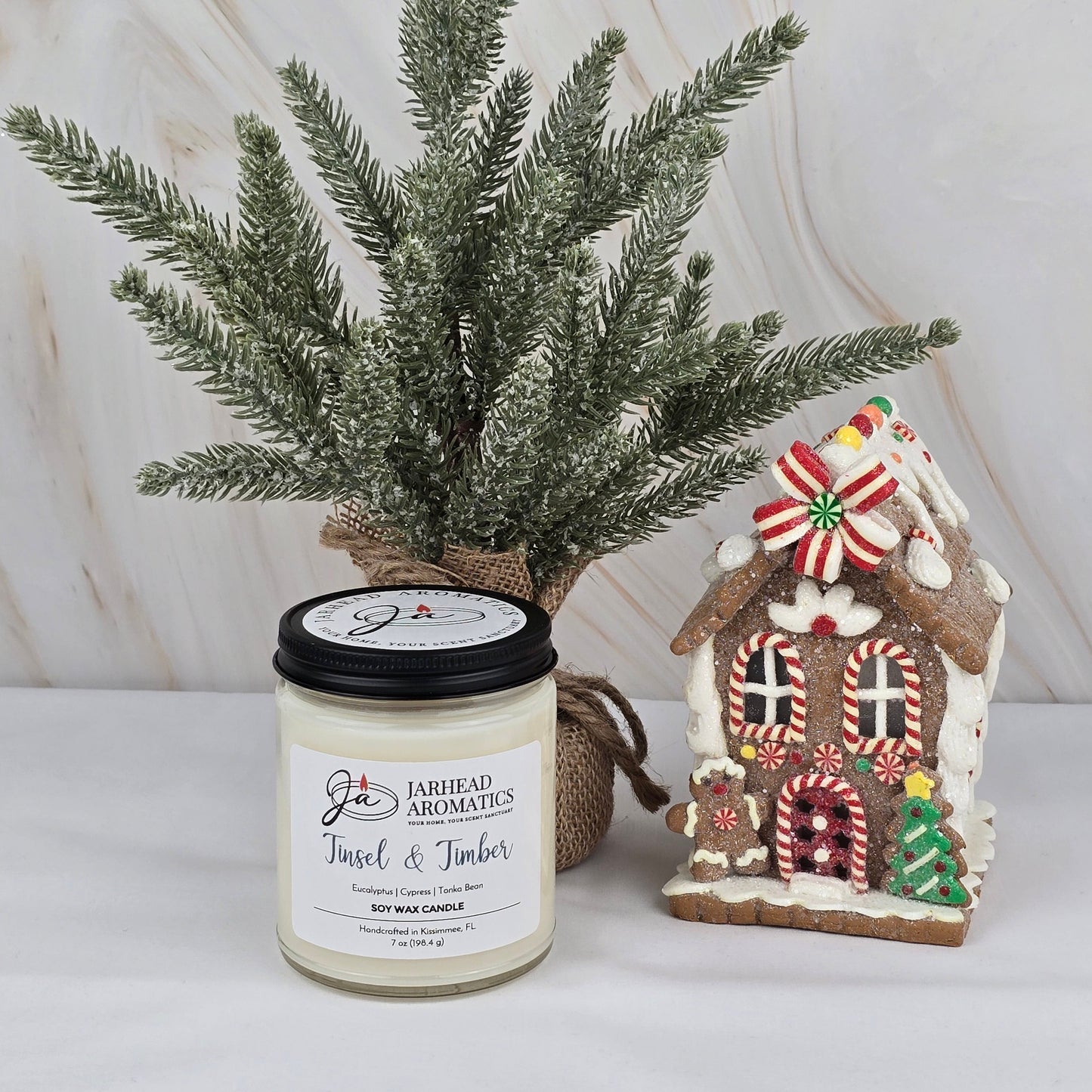 A festive scene features a small artificial pine tree, a decorated gingerbread house, and the Tinsel & Timber Soy Wax Candle from Jarhead Aromatics’ Winter Collection, all set against a marble background.