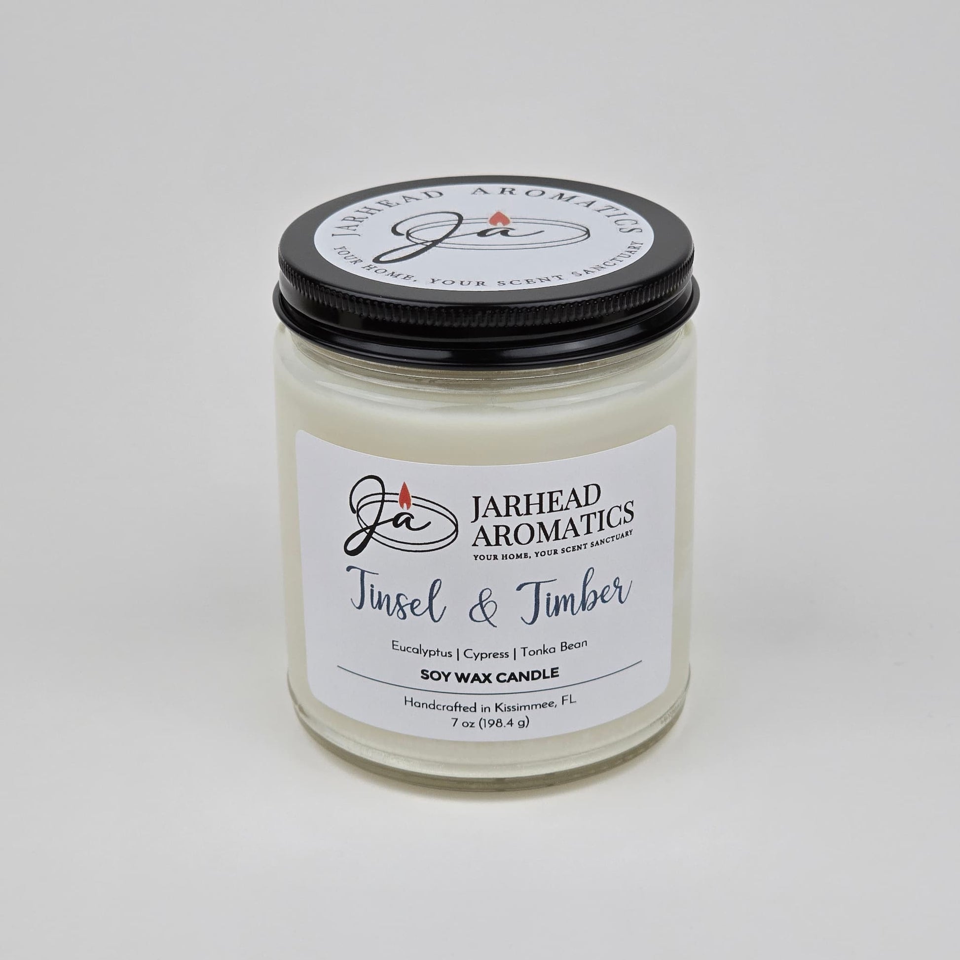 Introducing the Tinsel & Timber Soy Wax Candle by Jarhead Aromatics—a white soy wax candle in a clear jar with a black lid. Handcrafted in Kissimmee, FL, this holiday candle features an inviting blend of eucalyptus, pine, smoke, and tonka bean for a festive seasonal fragrance.