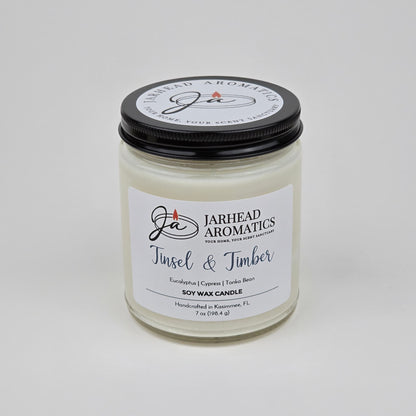 Introducing the Tinsel & Timber Soy Wax Candle by Jarhead Aromatics—a white soy wax candle in a clear jar with a black lid. Handcrafted in Kissimmee, FL, this holiday candle features an inviting blend of eucalyptus, pine, smoke, and tonka bean for a festive seasonal fragrance.