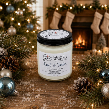 A Jarhead Aromatics Tinsel & Timber Soy Wax Candle, featuring festive notes of eucalyptus, pine, smoke, and tonka, sits on a snowy wooden table adorned with pine branches and ornaments in front of a glowing fireplace with stockings hanging above, filling the air with holiday cheer.