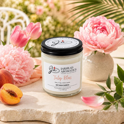 A Jarhead Aromatics Tulip Bliss Soy Wax Candle | Peach, Tulip & Soft Musk Candle rests on a table alongside pink peonies, a ripe peach, and green leaves. The bright, summery scene with a garden backdrop and gentle natural light perfectly highlights the fresh floral allure of this scented candle.