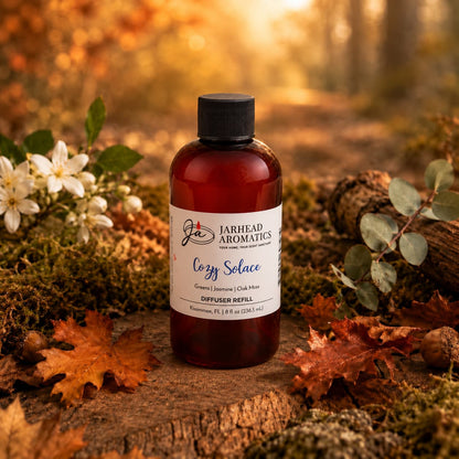 A bottle labeled Cozy Solace Diffuser Refill by Jarhead Aromatics stands on a forest path surrounded by autumn leaves, white flowers, and greenery, with warm sunlight filtering through the trees in the background—an inviting scene that perfectly captures the essence of this autumn home fragrance blend of fresh greens, jasmine, and oak moss.