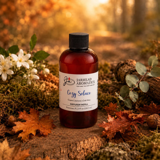 A bottle labeled Cozy Solace Diffuser Refill by Jarhead Aromatics stands on a forest path surrounded by autumn leaves, white flowers, and greenery, with warm sunlight filtering through the trees in the background—an inviting scene that perfectly captures the essence of this autumn home fragrance blend of fresh greens, jasmine, and oak moss.