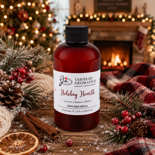 A brown plastic Holiday Hearth Diffuser Refill bottle by Jarhead Aromatics with a white label, centered on a rustic wooden table decorated with pine branches, cinnamon sticks, and dried orange slices in front of a blurred fireplace.