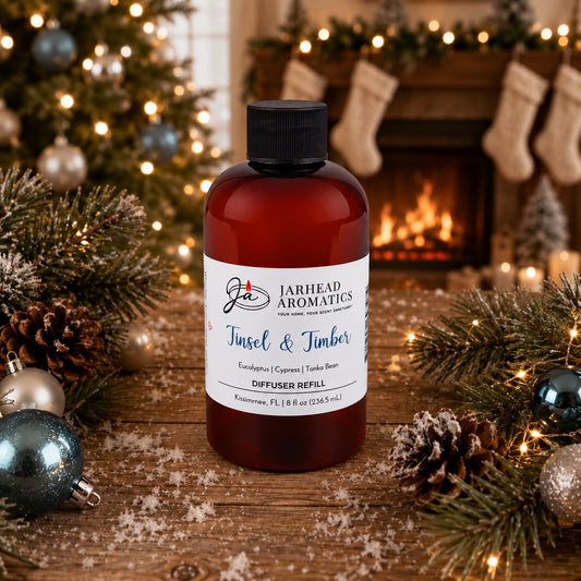Amber plastic bottle of Jarhead Aromatics Tinsel & Timber Diffuser Refill | Eucalyptus, Pine, Smoke & Tonka with a white label, sitting on a wooden table with pine branches, ornaments, and a fireplace in the background.