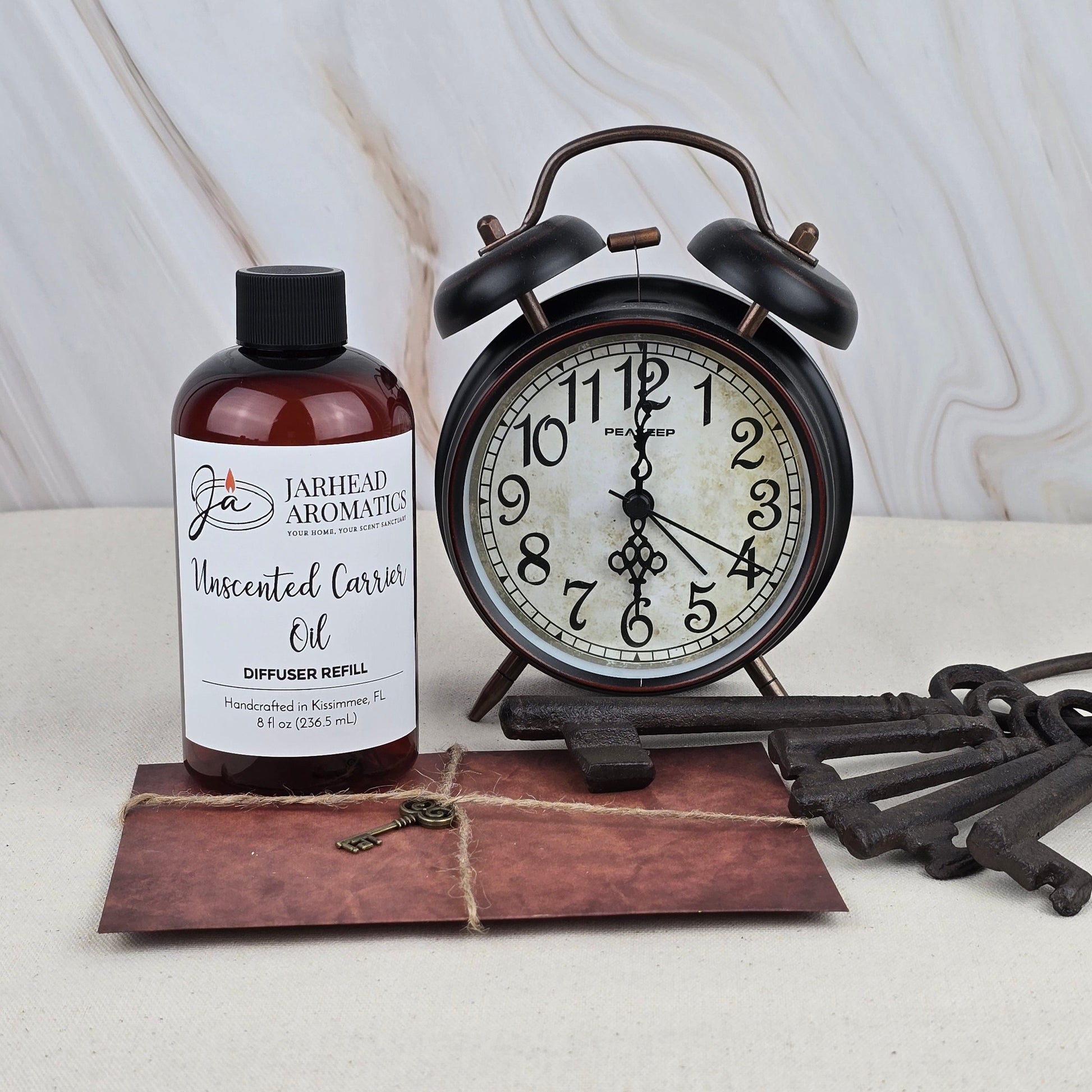 A bottle of Jarhead Aromatics Unscented Carrier Oil – Diffuser Refill sits beside a vintage black alarm clock, three antique keys, and a brown envelope on a light surface with a marble-patterned background.