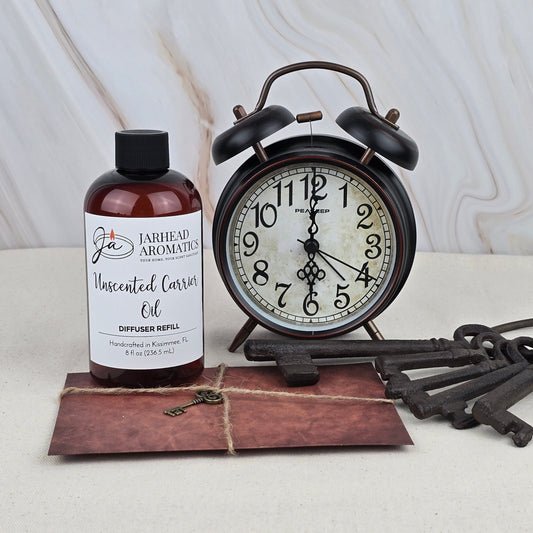 A bottle of Jarhead Aromatics Unscented Carrier Oil – Diffuser Refill sits beside a vintage black alarm clock, three antique keys, and a brown envelope on a light surface with a marble-patterned background.