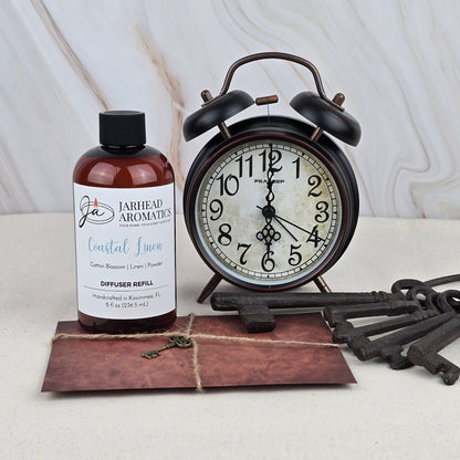 A bottle labeled Jarhead Aromatics Coastal Linen - Diffuser Refill, ideal for freshening rooms with a coastal scent, sits beside a vintage black alarm clock, five antique metal keys, and a twine-tied brown envelope on a light surface.