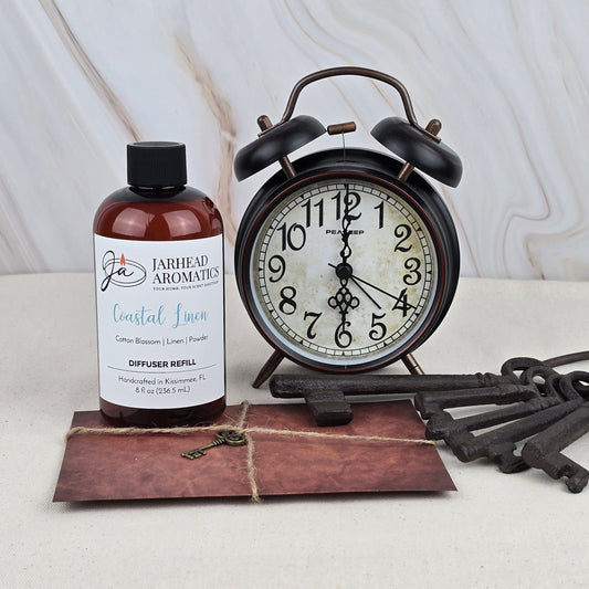 A bottle labeled Jarhead Aromatics Coastal Linen - Diffuser Refill, ideal for freshening rooms with a coastal scent, sits beside a vintage black alarm clock, five antique metal keys, and a twine-tied brown envelope on a light surface.