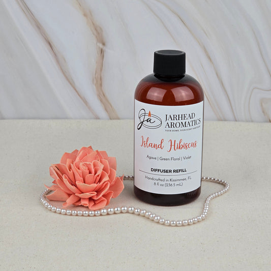 A bottle of Jarhead Aromatics Island Hibiscus - Diffuser Refill sits beside a coral artificial flower and pearls on a beige surface with a marble backdrop, evoking the vibe of a tropical paradise.