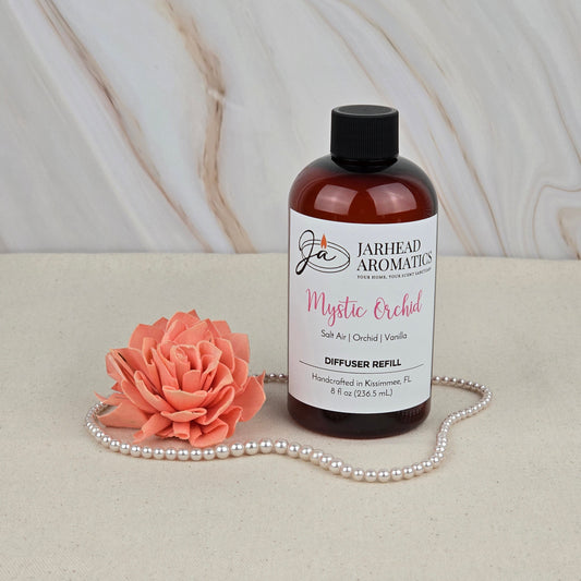 A bottle of Jarhead Aromatics Mystic Orchid - Diffuser Refill sits on a beige surface beside a peach artificial flower and pearls, evoking exotic orchid and coastal freshness with a marble-patterned backdrop.