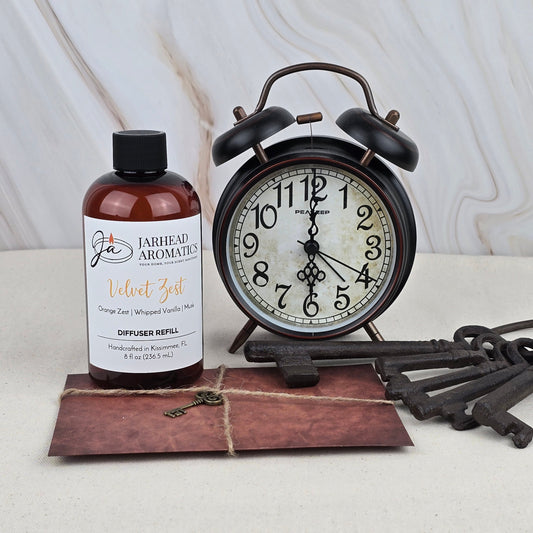 A brown bottle of Jarhead Aromatics Velvet Zest - Diffuser Refill stands beside a vintage black alarm clock and rustic keys atop an envelope, all arranged on a light wooden surface.