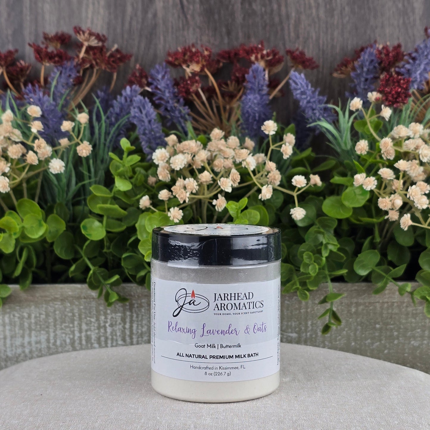 A jar of Jarhead Aromatics Lavender & Oats Milk Bath—Goat Milk & Buttermilk, All Natural—rests on a beige cushion, surrounded by green and purple artificial flowers with a rustic wood background.