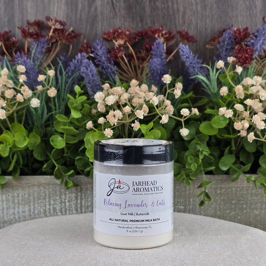 A jar of Jarhead Aromatics Lavender & Oats Milk Bath—Goat Milk & Buttermilk, All Natural—rests on a beige cushion, surrounded by green and purple artificial flowers with a rustic wood background.