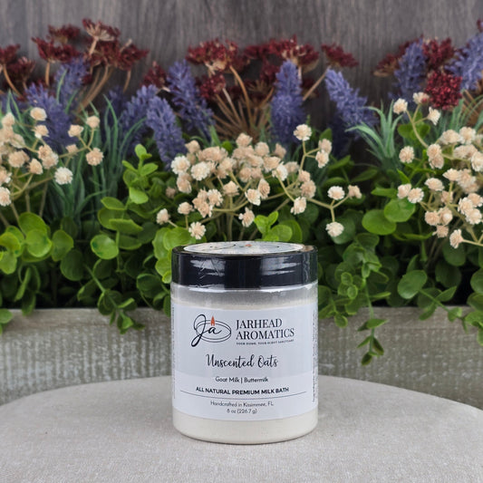 A jar of Jarhead Aromatics Unscented Oats Milk Bath - Goat Milk & Buttermilk - All Natural rests on a light surface, with green foliage and purple flowers in the background.