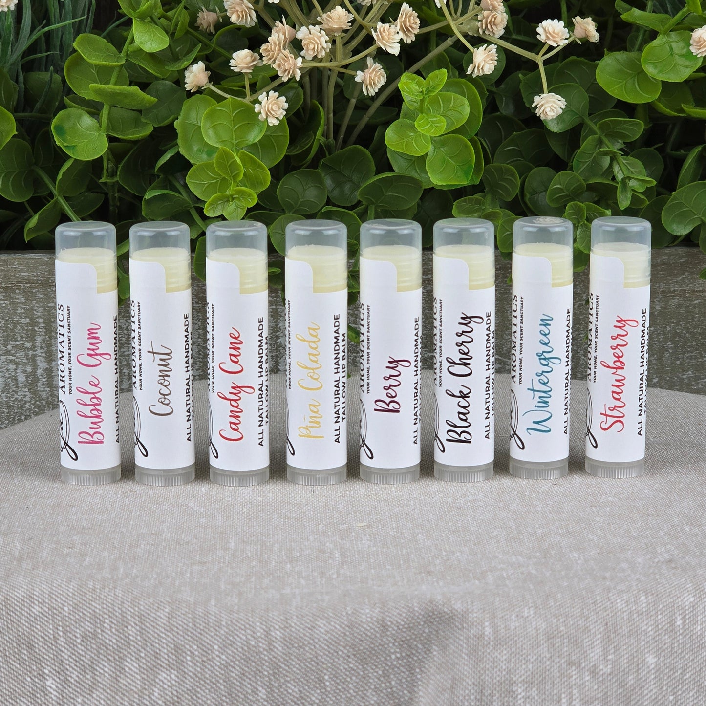 Eight Tallow Lip Balm - All Natural tubes by Jarhead Aromatics, each with a white label showing assorted flavors like Bubble Gum, Coconut, and Wintergreen, are arranged on a beige surface with green plants in the background.