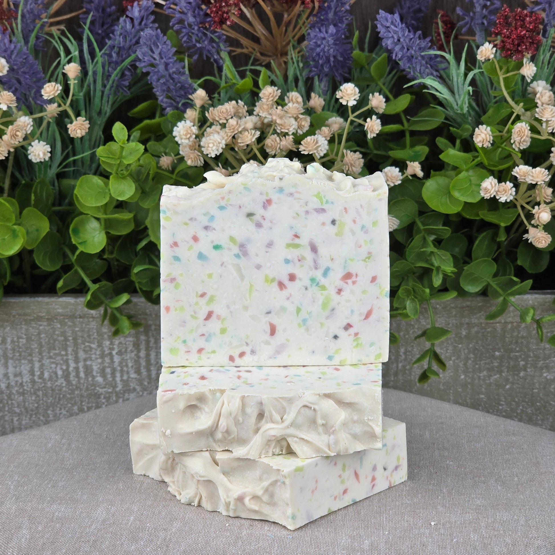Two bars of Jarhead Aromatics Almond + Berry Handmade Coconut Milk Artisan Soap, infused with shea butter and aloe, are stacked before vibrant artificial flowers and greenery on a neutral fabric surface.