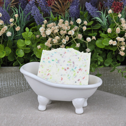 A bar of Jarhead Aromatics Almond + Berry Handmade Coconut Milk Artisan Soap stands upright in a white clawfoot bathtub dish, surrounded by green foliage and purple and red flowers.