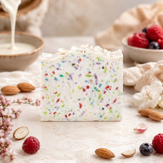 A vibrant Almond + Berry Handmade Coconut Milk Artisan Soap by Jarhead Aromatics stands on a marble surface, surrounded by almonds, berries, flowers, and a bowl of milk being poured in the background. Carefully crafted with sweet almond and juicy berry scents, this artisan soap brings a touch of luxury and freshness to your daily routine.