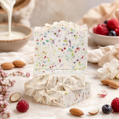Two bars of Jarhead Aromatics Almond + Berry Handmade Coconut Milk Artisan Soap, featuring colorful bits, rest on a light surface amidst almonds, berries, pink flowers, and a bowl of cream—creating a fresh, natural scene that highlights the charm of this sweet almond and juicy berry artisan soap.