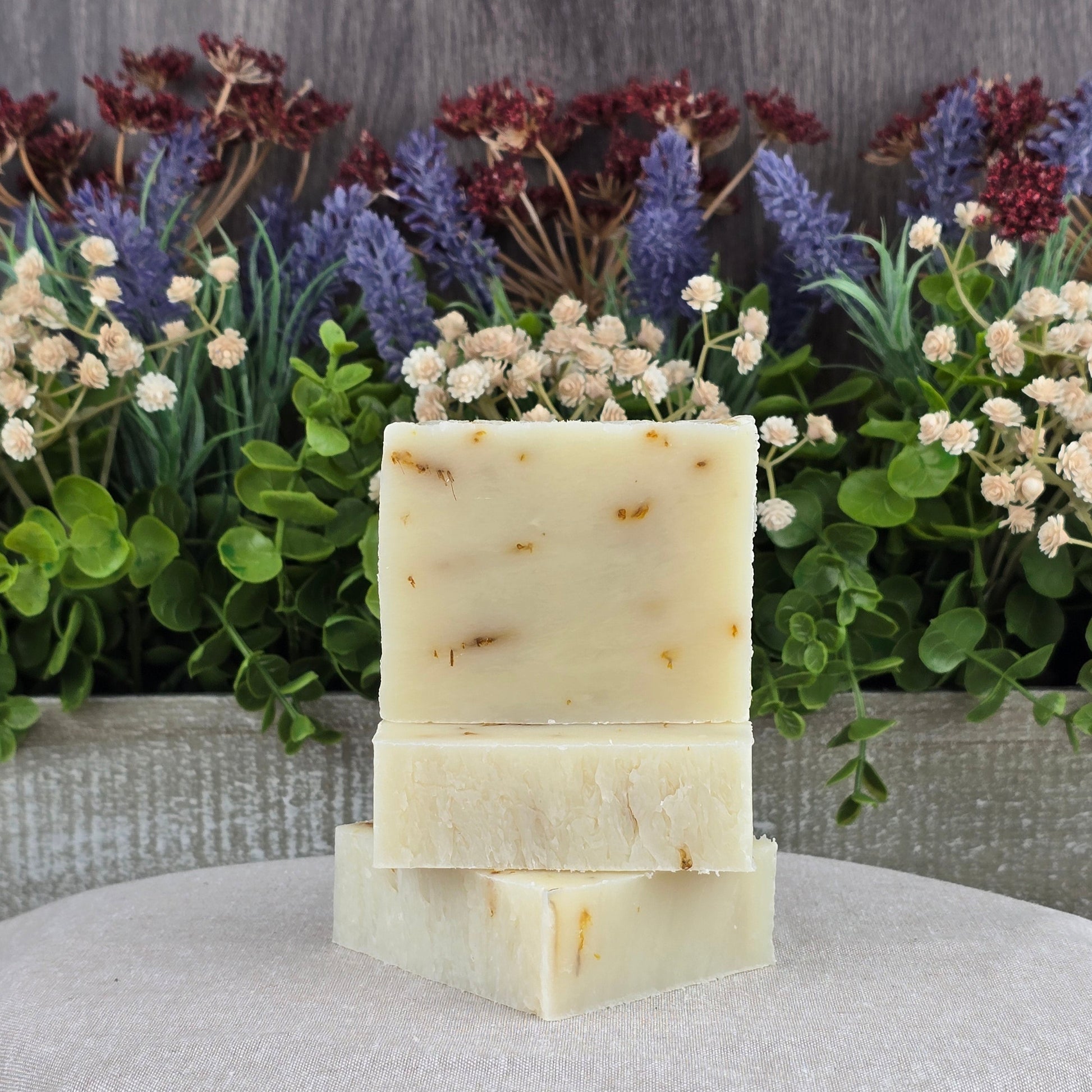 Three bars of Jarhead Aromatics Aloe + Calendula Handmade Soap, crafted with moisturizing oils, are stacked in front of green leaves and flowers on a wooden background. All natural and perfect for sensitive skin.