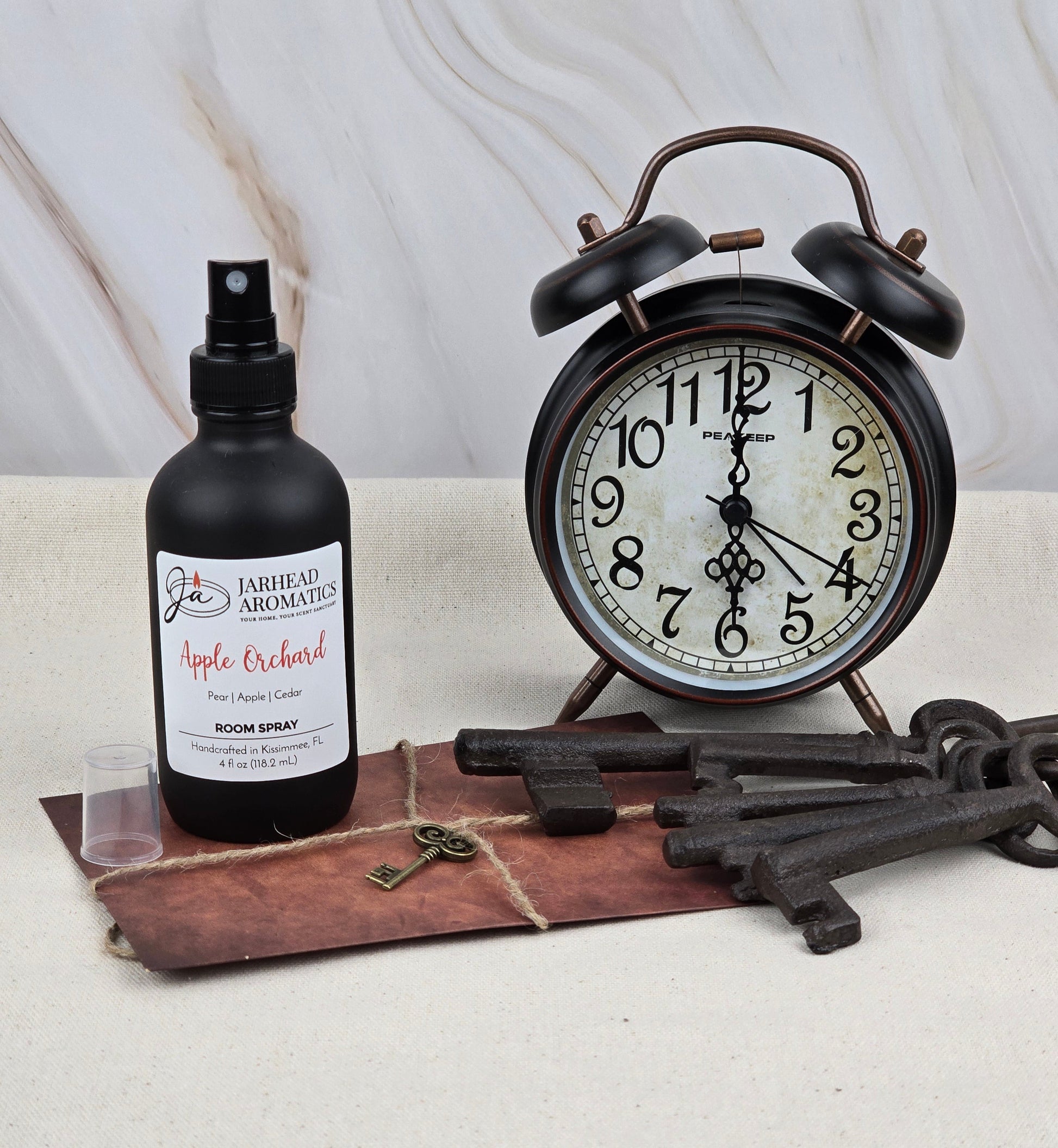 A vintage clock sits on a table beside Apple Orchard room spray by Jarhead Aromatics, with its fresh apple scent. Next to it, theres an antique key and a bunch of rustic keys on a leather mat, all against a marble-like backdrop.
