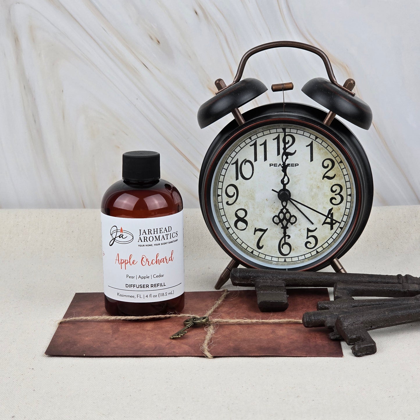 A bottle of Jarhead Aromatics Apple Orchard Diffuser Refill | Fresh Apple, Pear & Cedar sits next to a black alarm clock, three metal keys, and an envelope with a small key, all on a light surface—ready to fill your space with fresh fragrance.