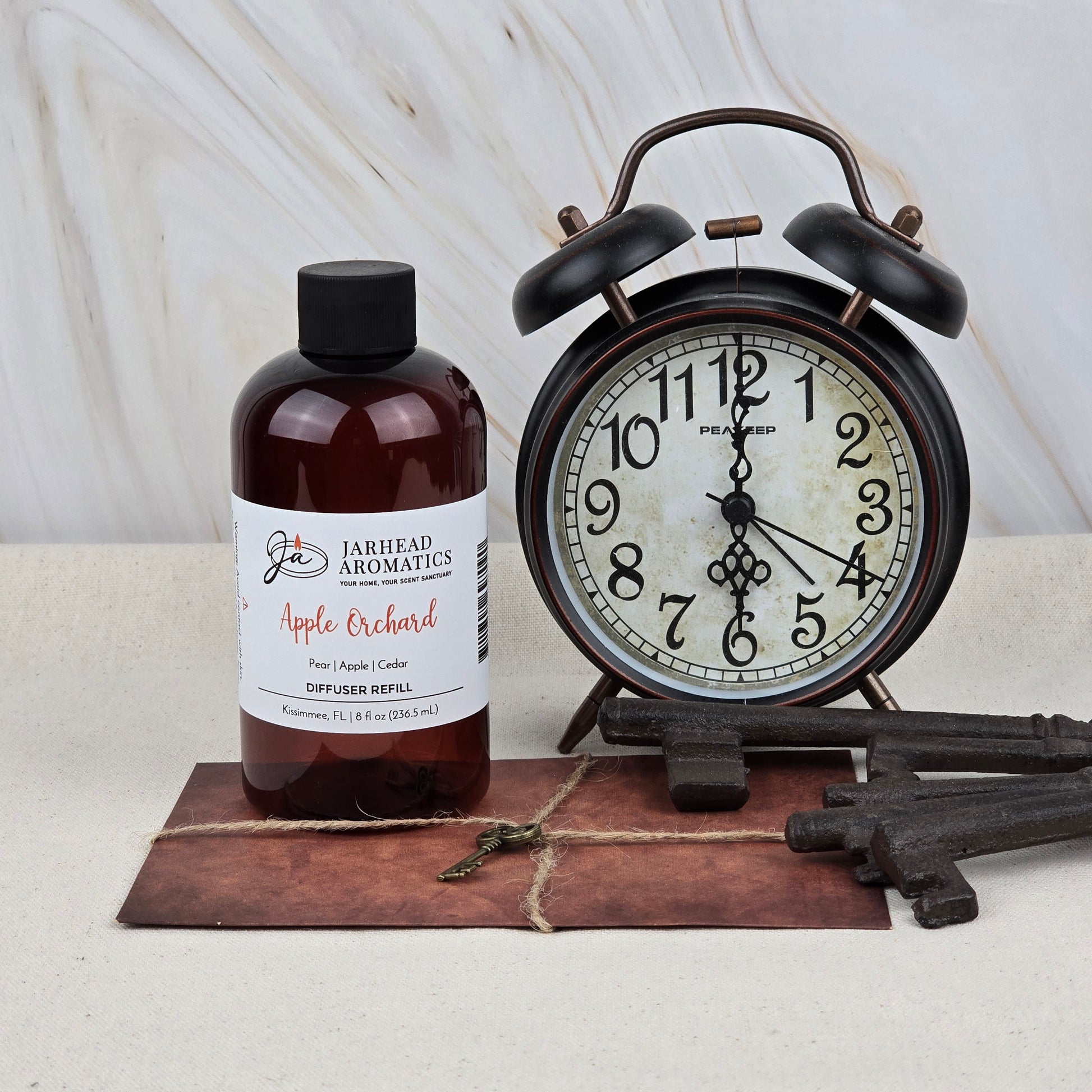 A bottle of Jarhead Aromatics Apple Orchard Diffuser Refill | Fresh Apple, Pear & Cedar stands beside a vintage black alarm clock showing 8:18, with old-fashioned keys and a rustic postcard on a light surface.