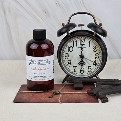 A bottle of Jarhead Aromatics Apple Orchard Diffuser Refill | Fresh Apple, Pear & Cedar stands beside a vintage black alarm clock showing 8:18, with old-fashioned keys and a rustic postcard on a light surface.