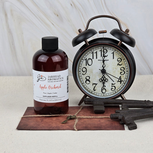 A bottle of Jarhead Aromatics Apple Orchard Diffuser Refill | Fresh Apple, Pear & Cedar stands beside a vintage black alarm clock showing 8:18, with old-fashioned keys and a rustic postcard on a light surface.
