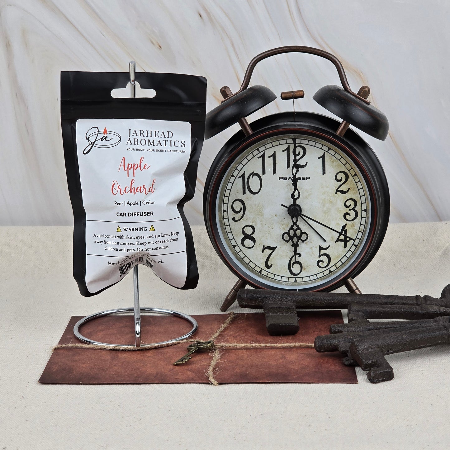A black alarm clock, vintage keys, and a Jarhead Aromatics Apple Orchard Car Diffuser | Fresh Crisp Apple Car Air Freshener are arranged on marble. The long-lasting scent packet hangs from a metal stand beside the clock.