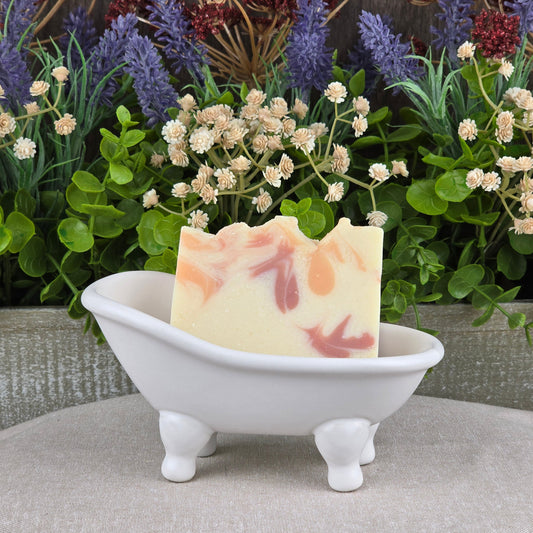 A decorative bar of Jarhead Aromatics’ Apple + Spice Handmade Coconut Milk Artisan Soap with a marbled pattern sits in a white clawfoot bathtub dish, accented by green leaves and purple and white artificial flowers.