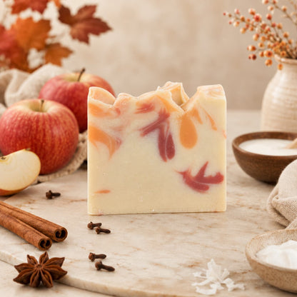 A bar of Jarhead Aromatics Apple + Spice Handmade Coconut Milk Artisan Soap, featuring orange and red swirls, rests on a marble surface surrounded by apples, cinnamon sticks, star anise, cloves, a bowl of powder, dried leaves, and a rustic vase filled with orange berries. This artisan Crisp Apple & Warm Spice Soap fills your home with the comforting aroma of apple spice.