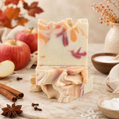 Two artisanal bars of Jarhead Aromatics’ Apple + Spice Handmade Coconut Milk Artisan Soap, featuring a beautiful swirl pattern, are stacked on a marble surface amid fresh apples, cinnamon sticks, star anise, a wooden bowl of fragrant powder, and scattered autumn leaves. These Crisp Apple & Warm Spice soaps embody the cozy essence of the season with their inviting fragrance and expert craftsmanship.