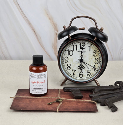 A vintage alarm clock with Roman numerals is next to a Jarhead Aromatics Apple Orchard Reed Diffuser Refill, filling the air with fresh apple scents. They sit on a light surface with large old-fashioned keys and a twine-tied envelope against a muted marble background.