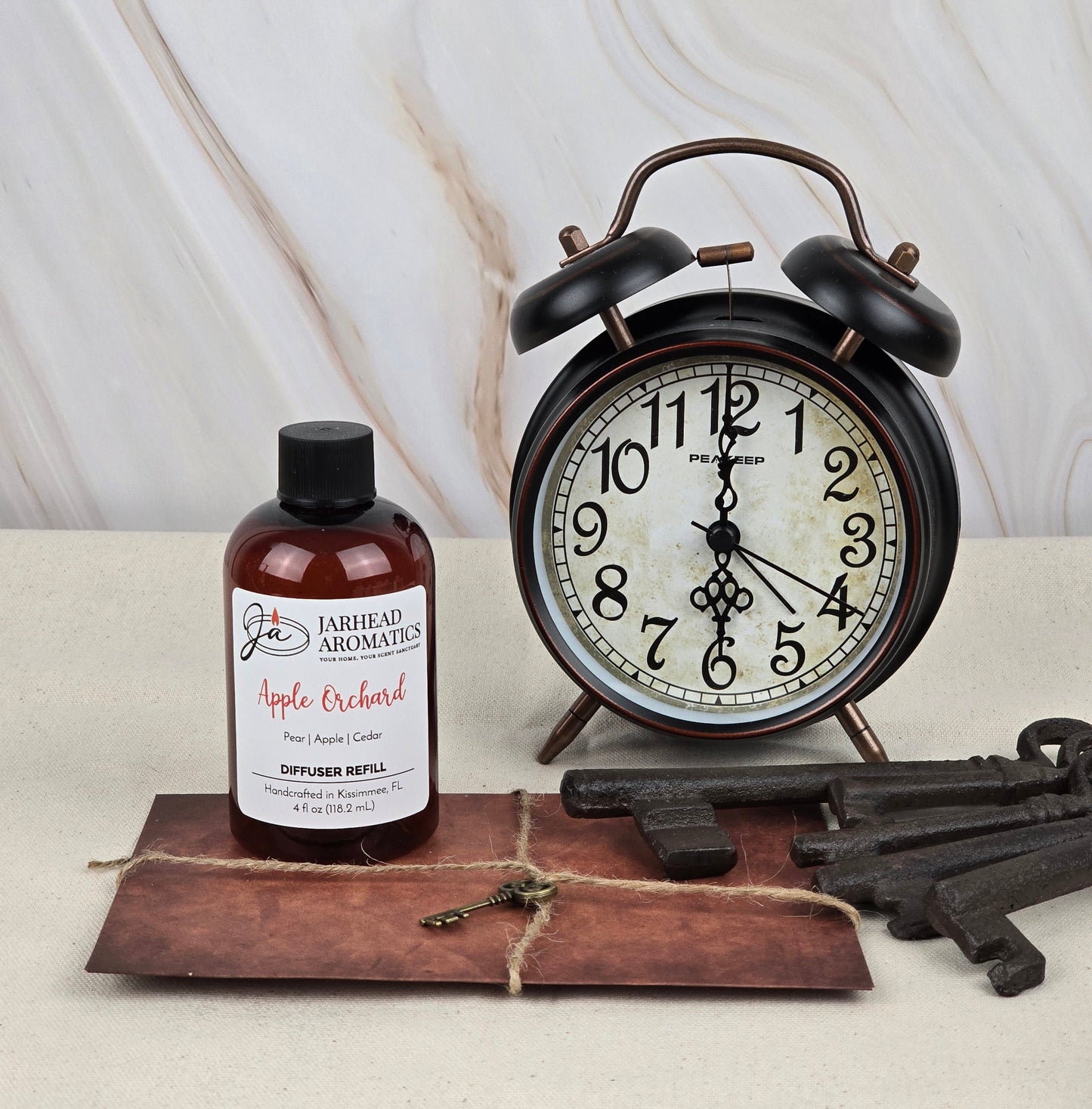 A Jarhead Aromatics Apple Orchard Reed Diffuser Refill, exuding fresh apple fragrance, is on a table next to a vintage alarm clock. Large metal keys lie on a brown envelope nearby, against a marble-patterned wall.