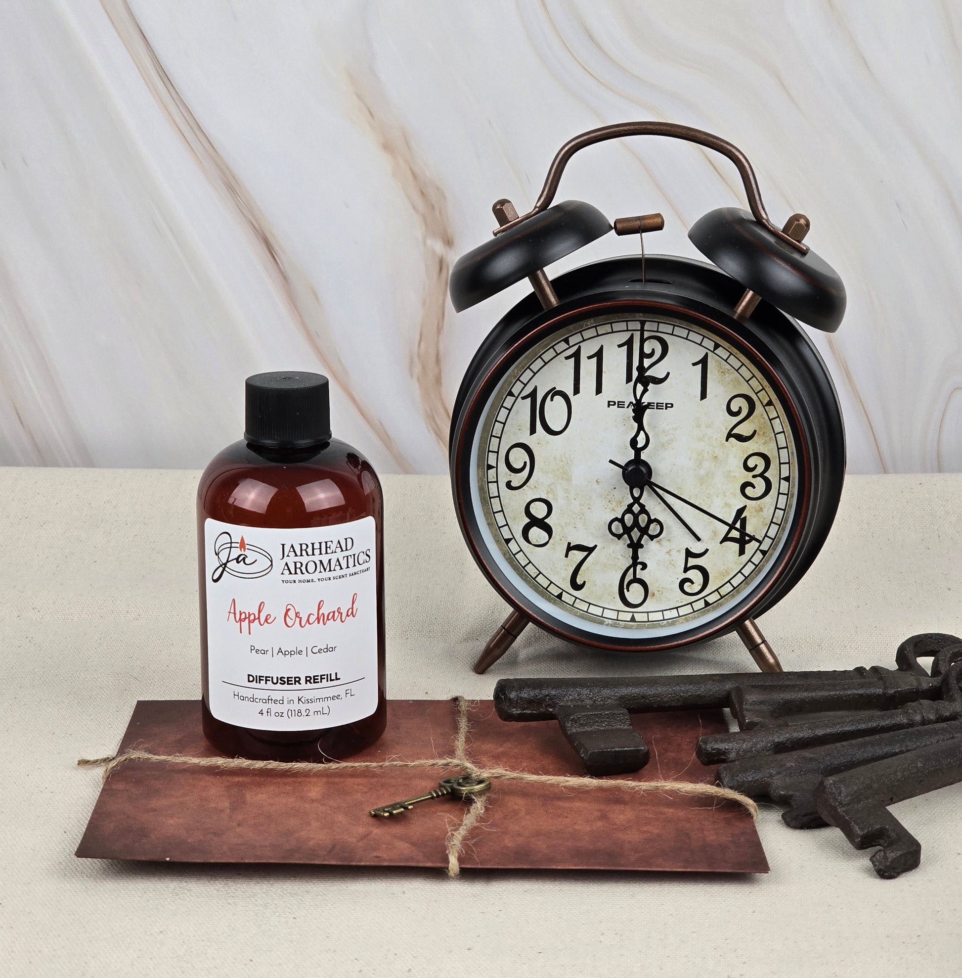 A Jarhead Aromatics Apple Orchard Reed Diffuser Refill, exuding fresh apple fragrance, is on a table next to a vintage alarm clock. Large metal keys lie on a brown envelope nearby, against a marble-patterned wall.