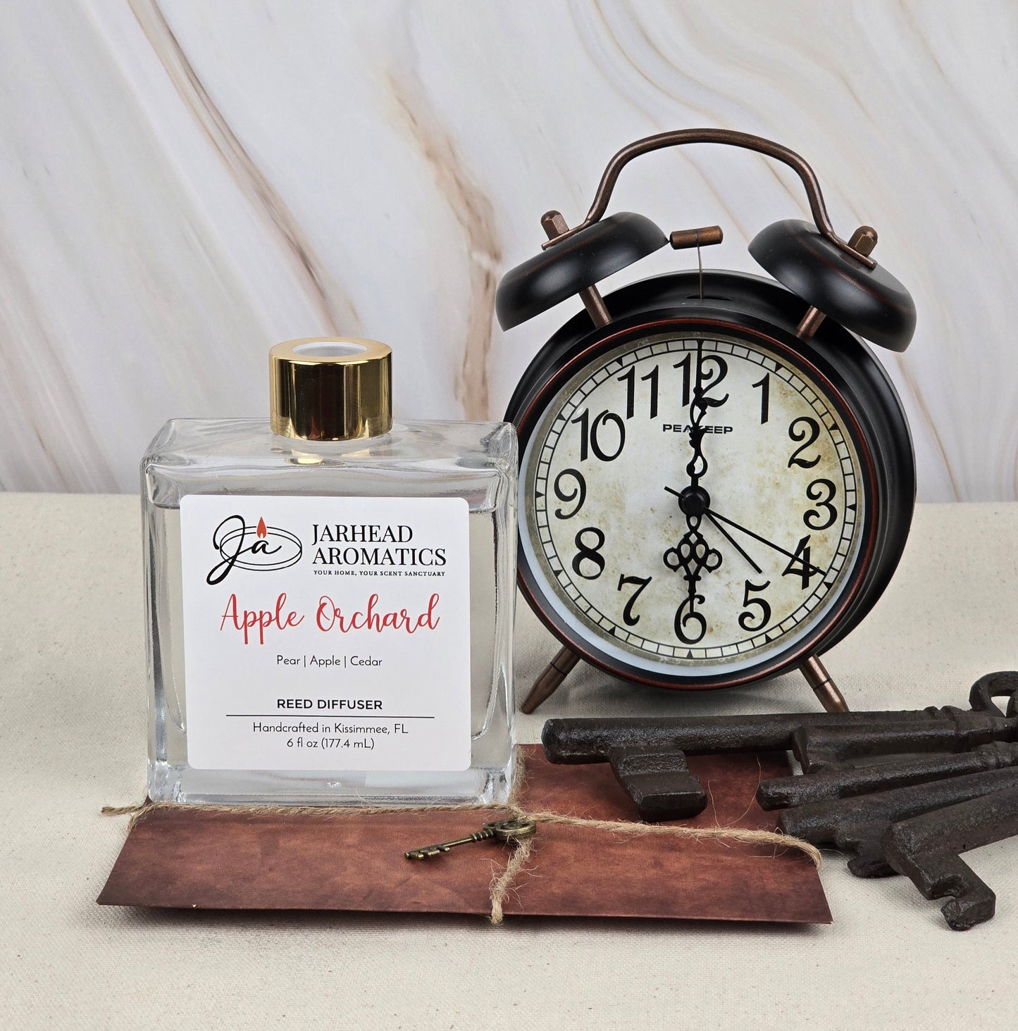 The Apple Orchard Reed Diffuser by Jarhead Aromatics, infused with apple-scented essential oils, stands beside a vintage-style alarm clock. Three skeleton keys rest on a brown leather tag in the foreground, set against an elegant marble surface backdrop.