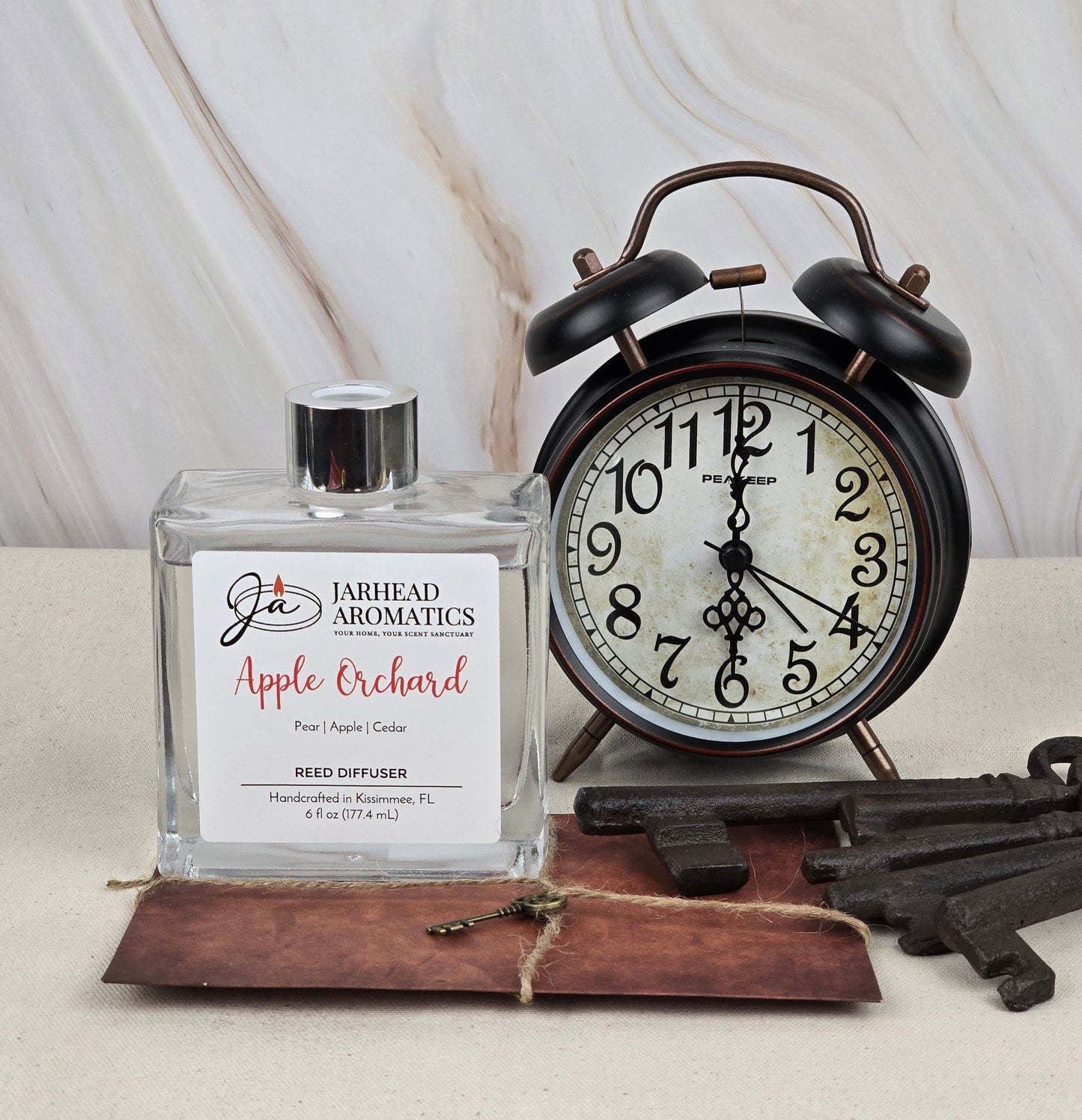 A vintage-style alarm clock shows 10:10 beside a square bottle labeled Apple Orchard - Reed Diffuser by Jarhead Aromatics, offering apple, pear, and cedar aromas. Three old-fashioned keys lie on a brown leather surface nearby, evoking nostalgia with each glance.