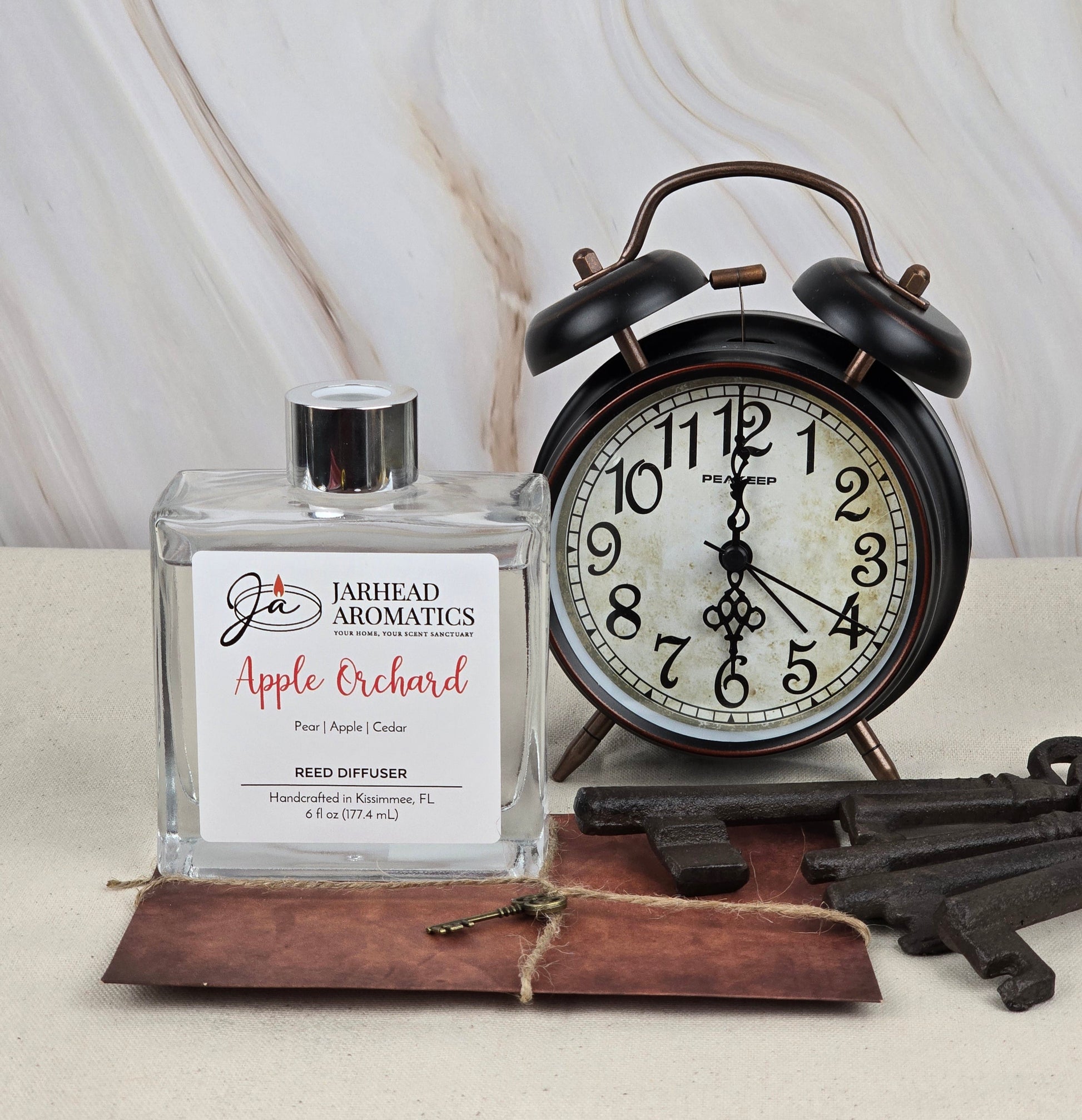 A vintage-style alarm clock shows 10:10 beside a square bottle labeled Apple Orchard - Reed Diffuser by Jarhead Aromatics, offering apple, pear, and cedar aromas. Three old-fashioned keys lie on a brown leather surface nearby, evoking nostalgia with each glance.