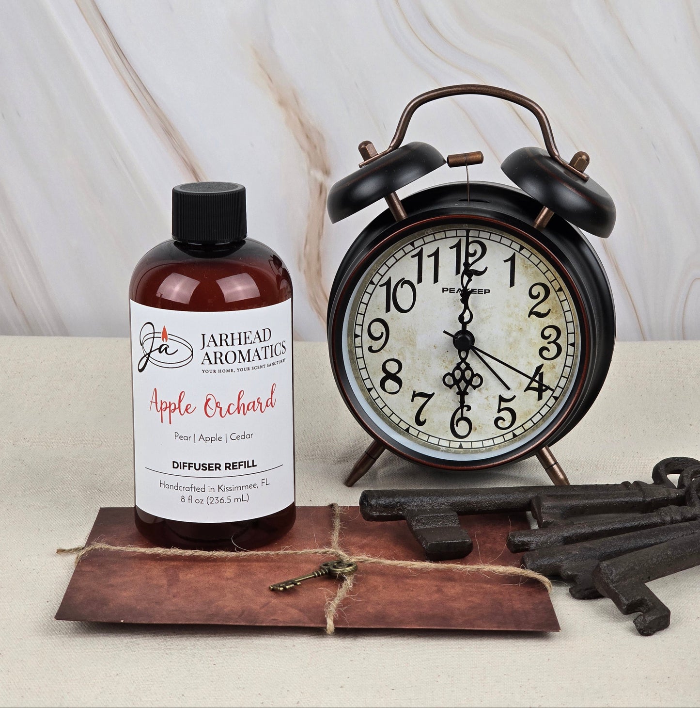 A Jarhead Aromatics Apple Orchard Reed Diffuser Refill, emitting a fresh apple scent, sits beside a vintage black alarm clock set to 10:10. Nearby, rustic metal keys lie on a brown leather pouch against a marble background.