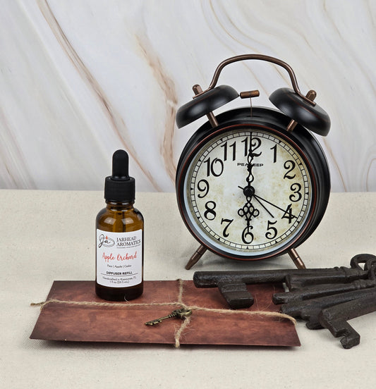 A vintage alarm clock is on a table next to a bottle labeled Apple Orchard by Jarhead Aromatics, exuding a fresh apple scent, with an envelope tied with string and three antique keys nearby. The background features a white and brown marble pattern.