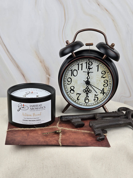 A black vintage alarm clock shows 10:10 beside a Jarhead Aromatics Artisan Bread Coconut Wax Blend Candle on a brown envelope with twine and antique keys, all set against a marble-patterned background.