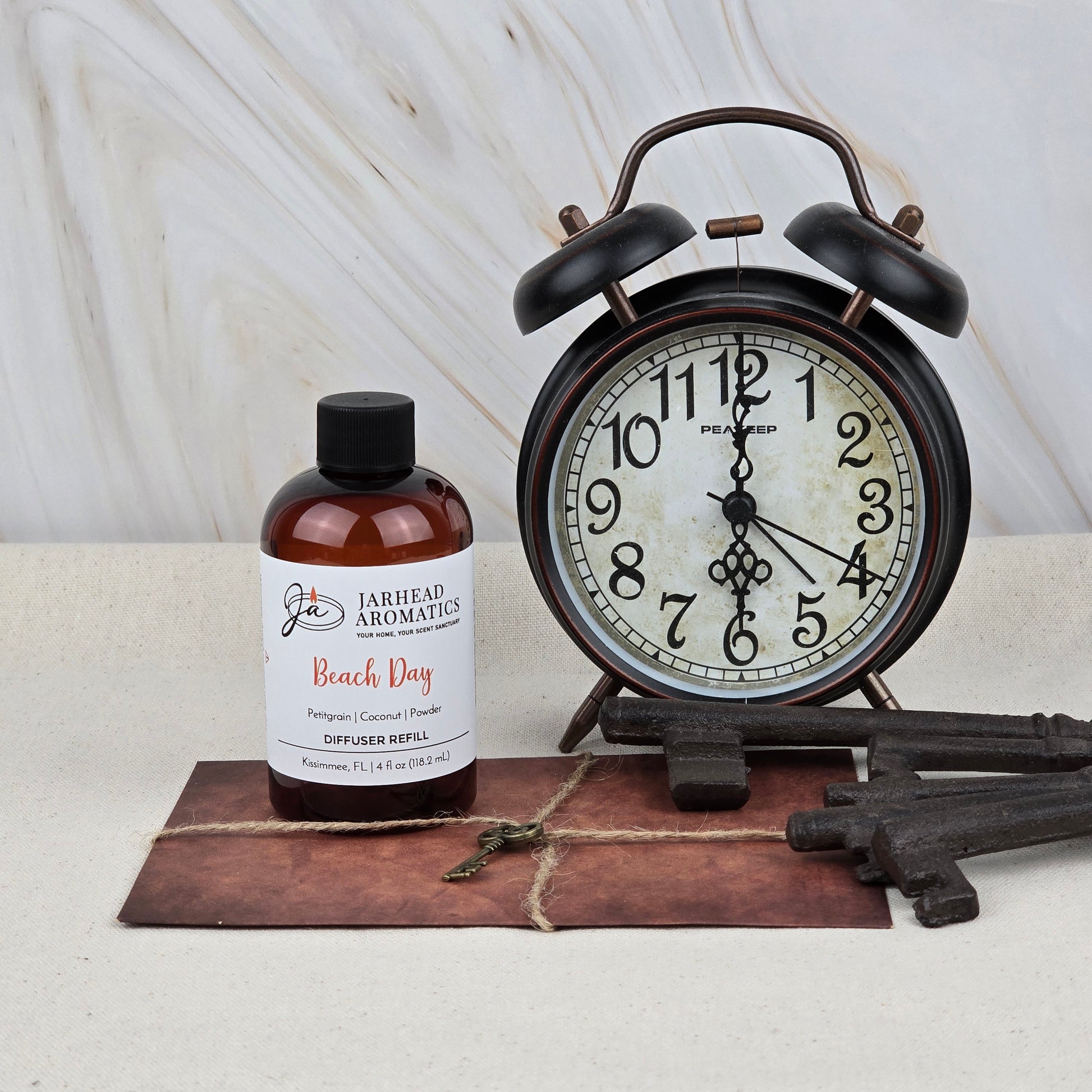 A vintage black alarm clock, three antique keys, and a Jarhead Aromatics Beach Day Diffuser Refill (Coconut, Citrus & Cotton Blossom) are arranged on a brown envelope atop light marble, evoking a fresh coastal scent.