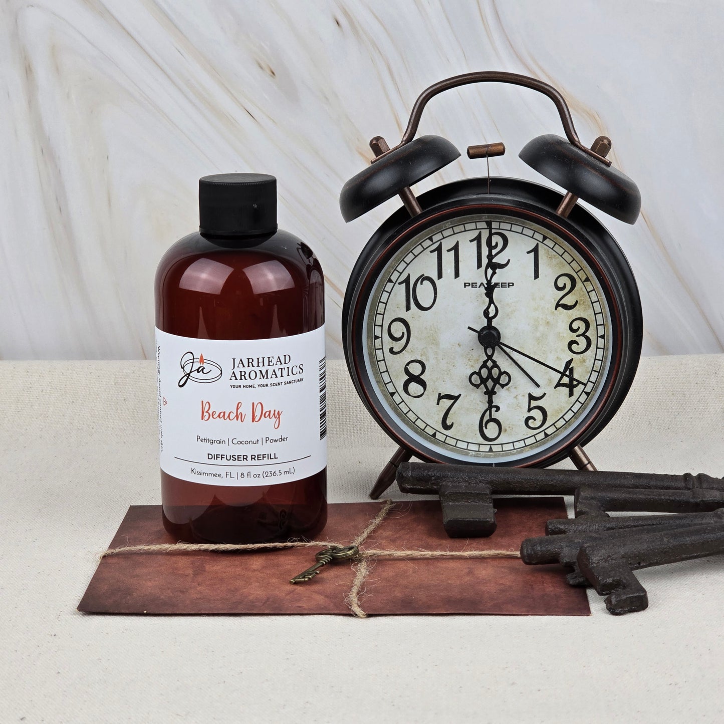 A bottle of Jarhead Aromatics Beach Day Diffuser Refill | Coconut, Citrus & Cotton Blossom sits beside a vintage black alarm clock, three antique keys, and an old postcard on a light surface with a marble background.